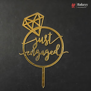 A gold metal 'just engaged' cake topper with a diamond graphic and stake, displayed on a dark textured background. A Bakeyy logo is visible in the top right corner.