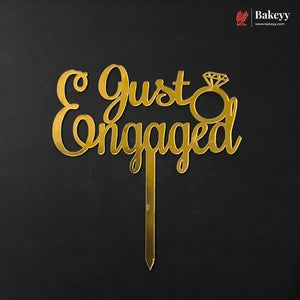 Gold laser-cut acrylic cake topper with the words 'Just Engaged' and a ring graphic, shown on a black background with a Bakeey logo in the top right corner.
