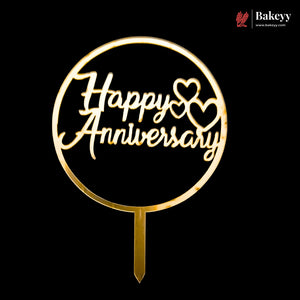 A gold-colored metal cake topper with a circular frame and stake, featuring the words 'Happy Anniversary' in cursive script with two heart shapes, shown against a solid black background. A 'Bakeyy' brand watermark is visible in the top right corner.