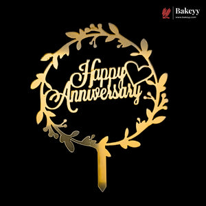 A gold-colored wooden cake topper with a circular leaf wreath design and the words 'Happy Anniversary' in cursive script, shown on a solid black background with a Bakeey brand watermark in the top right corner.