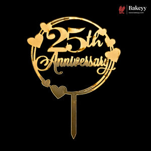 A gold-colored metal cake topper featuring the text '25th Anniversary' inside a circular frame with heart accents, shown on a solid black background. A Bakeyy brand logo is visible in the top right corner.