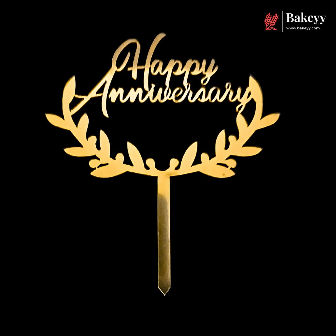 Gold laser-cut acrylic cake topper with 'Happy Anniversary' script and leafy wreath design on a black background, with a Bakeyy logo in the top right corner.