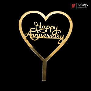A gold heart-shaped cake topper with 'Happy Anniversary' written in elegant cursive script, set against a solid black background. A Bakeey brand logo is visible in the top right corner.