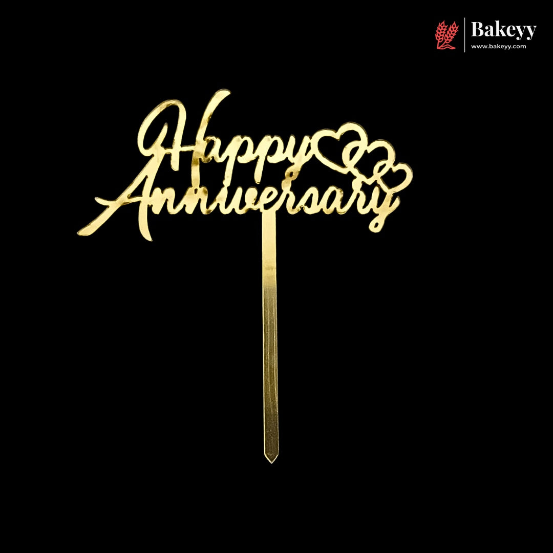 Gold metal 'Happy Anniversary' cake topper with cursive lettering and heart details on a black background, with a Bakeyy logo in the top right corner.