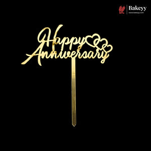 Gold metal 'Happy Anniversary' cake topper with cursive lettering and heart details on a black background, with a Bakeyy logo in the top right corner.