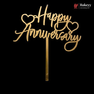 Gold laser-cut acrylic 'Happy Anniversary' cake topper with heart details and a metal stand, shown against a solid black background. A Bakeyy brand logo is visible in the top right corner.