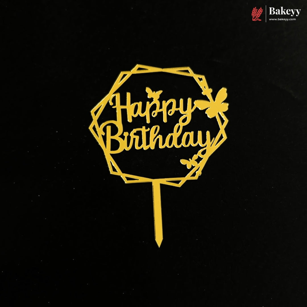 A gold metal 'Happy Birthday' cake topper with a geometric octagonal frame, decorative butterflies, and a stick, shown on a black background. A 'Bakeey' brand logo is visible in the top right corner.
