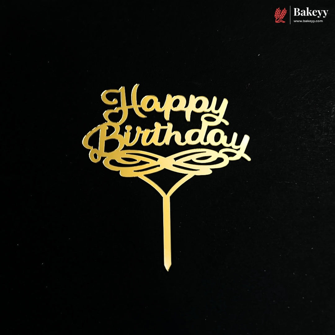 A gold metal 'Happy Birthday' cake topper with elegant cursive lettering and an ornate base, shown on a solid black background. A Bakeyy brand logo is visible in the top right corner.