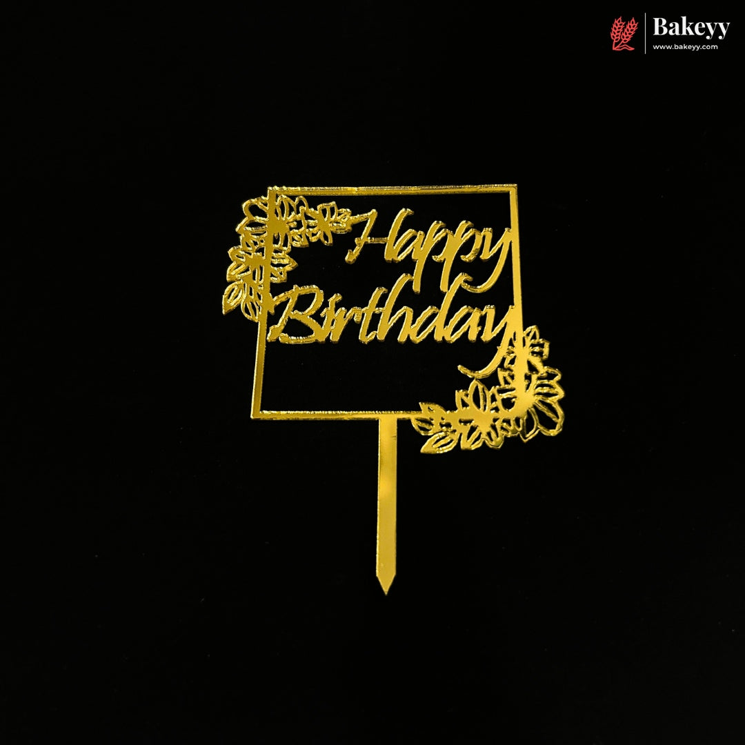 A gold metal 'Happy Birthday' cake topper with decorative floral cutouts and a stake, shown against a solid black background. A Bakeyy watermark is visible in the top right corner.