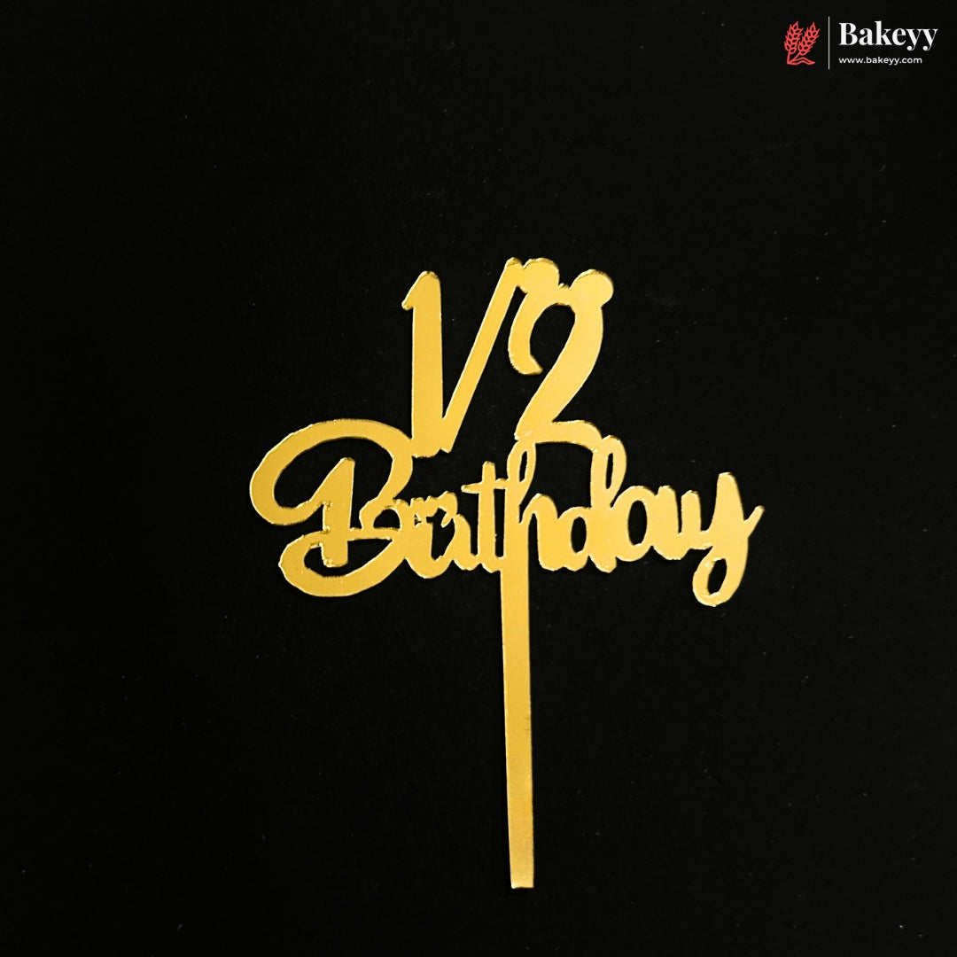 Gold metal cake topper with '1/2 Birthday' script on a black background, featuring a Bakeyy brand logo in the top right corner.