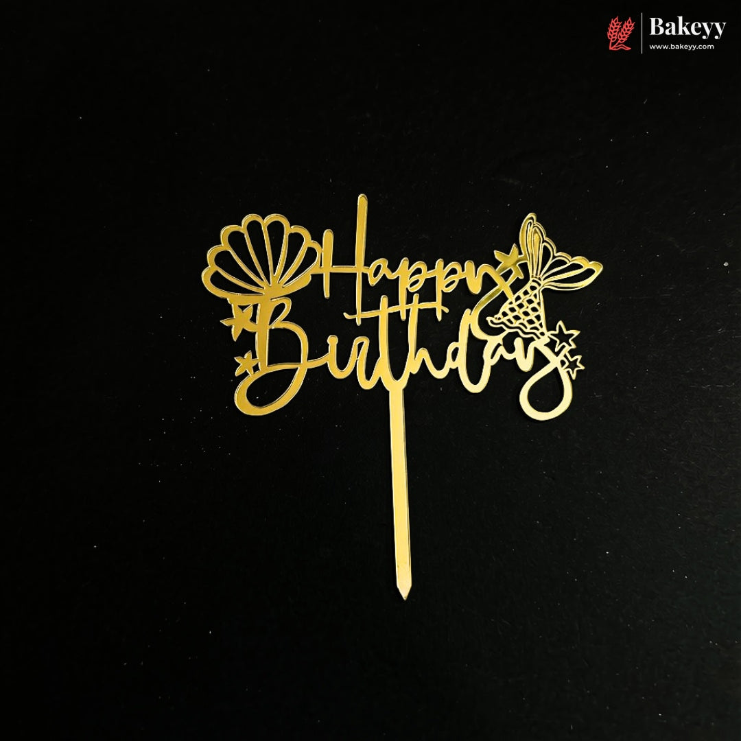 A gold metal cake topper with 'Happy Birthday' script, decorated with a seashell and butterfly design, shown on a black background with a Bakeyy brand watermark in the top right corner.