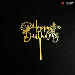 A gold metal cake topper with 'Happy Birthday' script, decorated with a seashell and butterfly design, shown on a black background with a Bakeyy brand watermark in the top right corner.