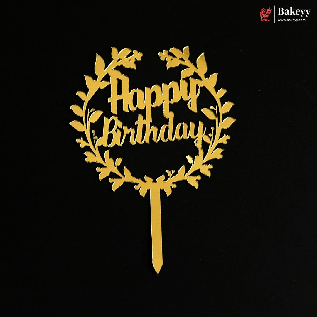 A gold metallic 'Happy Birthday' cake topper with a leafy wreath design and a straight stake, shown on a solid black background. A Bakeey brand logo is visible in the top right corner.
