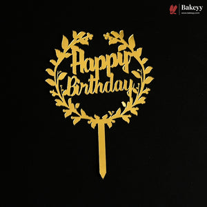 A gold metallic 'Happy Birthday' cake topper with a leafy wreath design and a straight stake, shown on a solid black background. A Bakeey brand logo is visible in the top right corner.