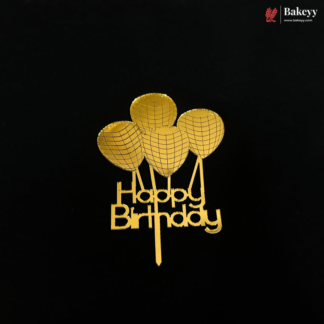 A gold-colored metal cake topper featuring three balloon shapes above the words 'Happy Birthday' in cursive, displayed on a solid black background. A 'Bakeyy' brand logo is visible in the top right corner.