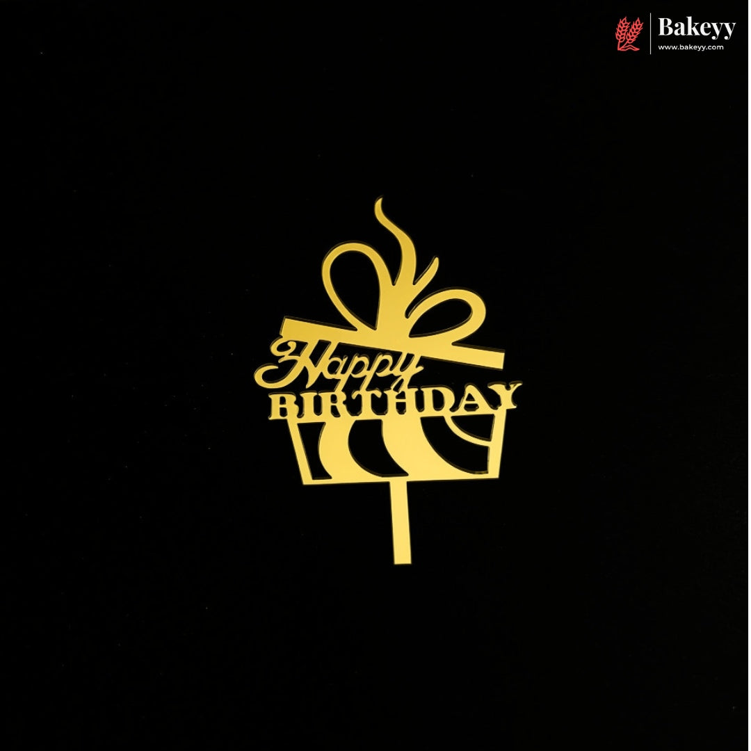 A gold metal cake topper shaped like a gift box with a bow and the words 'Happy Birthday' on a black background. A small Bakeey brand logo is visible in the top right corner.