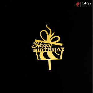 A gold metal cake topper shaped like a gift box with a bow and the words 'Happy Birthday' on a black background. A small Bakeey brand logo is visible in the top right corner.