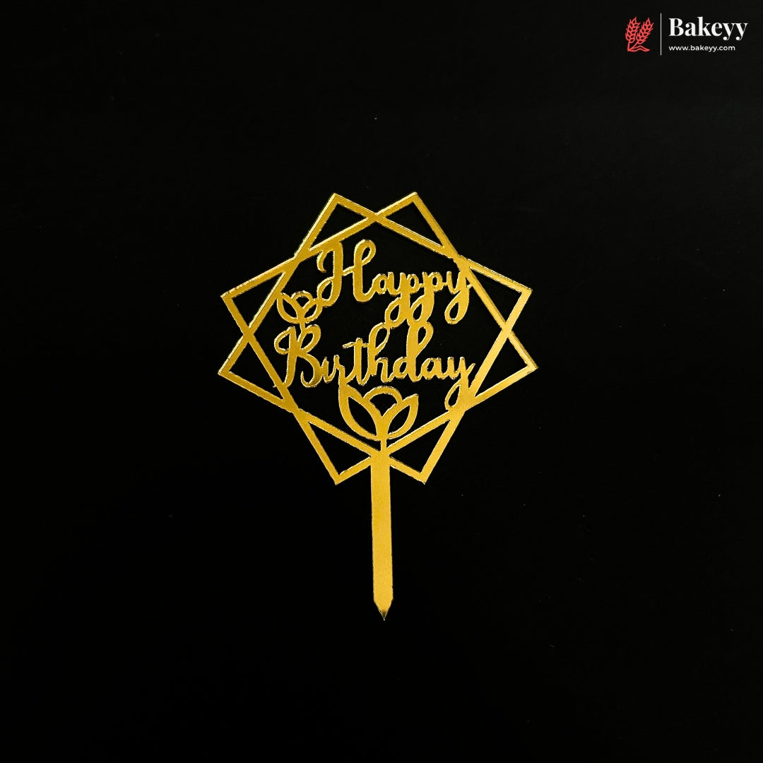 A gold metal cake topper with a geometric diamond frame and the words 'Happy Birthday' in cursive, displayed on a solid black background. A Bakeyy logo is visible in the top right corner.