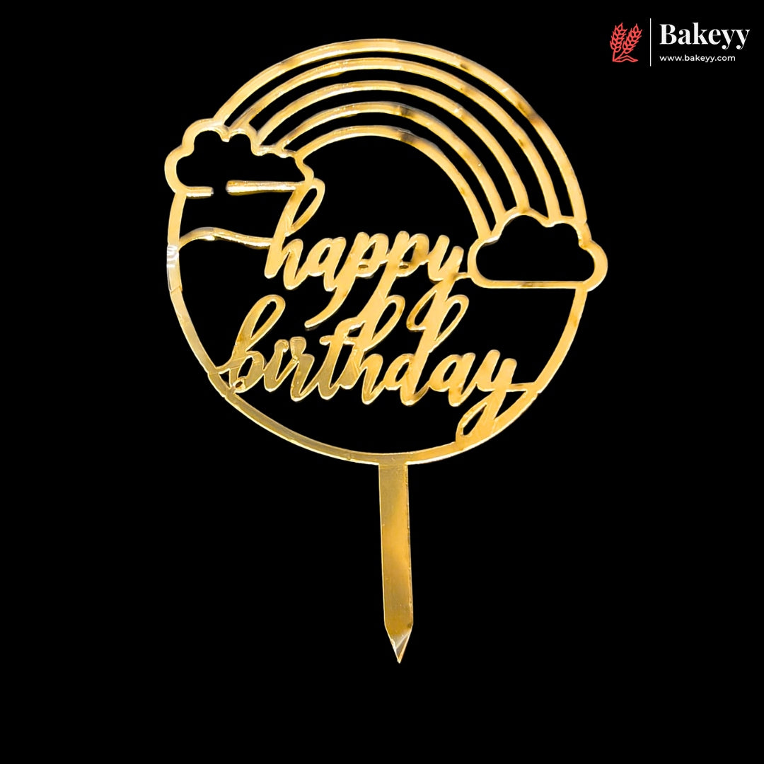 A gold-colored acrylic 'Happy Birthday' cake topper with a rainbow, clouds, and stick, shown on a solid black background. A 'Bakeey' brand logo is visible in the top right corner.