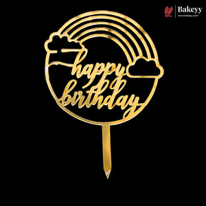 A gold-colored acrylic 'Happy Birthday' cake topper with a rainbow, clouds, and stick, shown on a solid black background. A 'Bakeey' brand logo is visible in the top right corner.