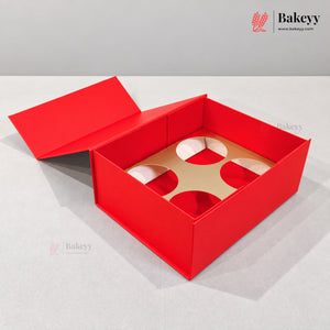 A red bakery gift box with a hinged lid open, showing a tan cardboard insert with four circular cutouts, photographed on a light gray surface against a gray background. The Bakeyy logo appears in the top right corner.