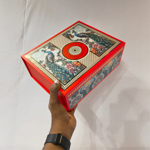 A hand holds a decorative red rectangular box with peacock illustrations and a circular red-and-gold lid emblem, photographed against a plain light background.