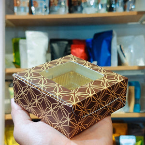 A hand holds a square brown gift box with a clear window and a geometric gold foil pattern, set against a blurred background of shelves with packaged goods.