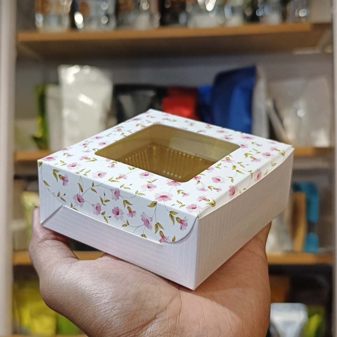 A hand holding a square white cardboard box with a floral pattern and a clear window showing a gold-colored insert tray inside, set against a blurred background of shelves.