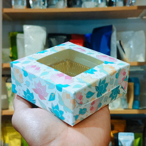 A hand holds a square floral-patterned gift box with a clear window showing a gold foil insert, set against a blurred background of shelves and packaged items.