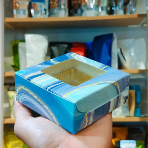 A hand holding a small square gift box with a marbled blue, white, and gold pattern and a clear window showing a gold foil insert inside, set against a blurred background of shelves with packaged goods.