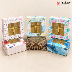 Five decorative bakery boxes with clear plastic window tops, arranged on a beige studio background. The boxes feature floral, striped, geometric gold patterns, and a blue marbled design.