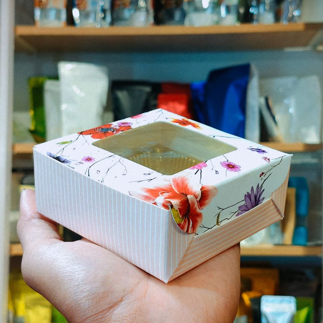 A hand holding a square white paper gift box with a clear window and floral printed edges, set against a blurred background of shelves with packaged items.