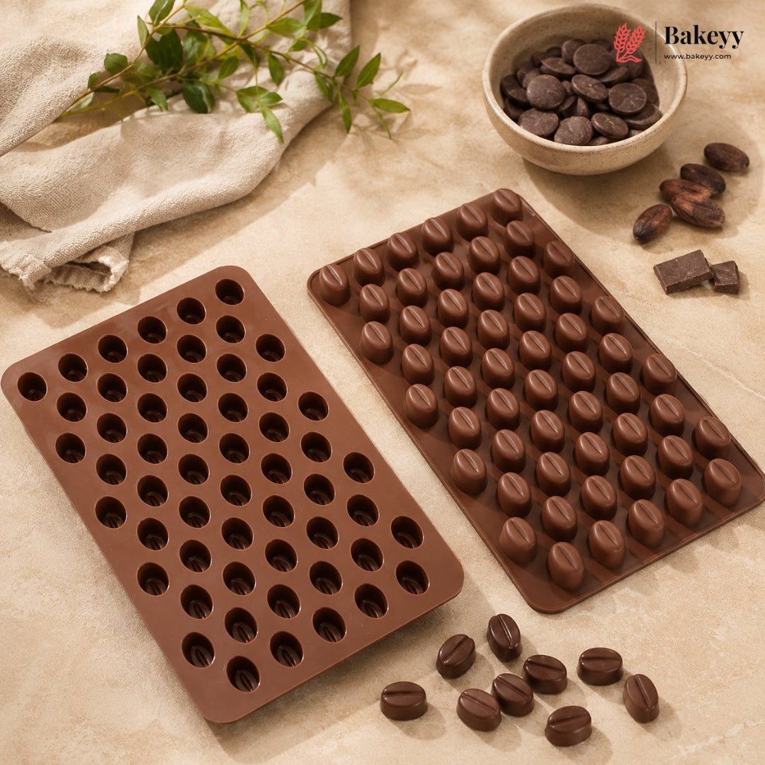 Two brown silicone molds for making coffee bean-shaped chocolates, displayed on a beige textured surface with chocolate pieces, a bowl of chocolate chips, and a small plant sprig.