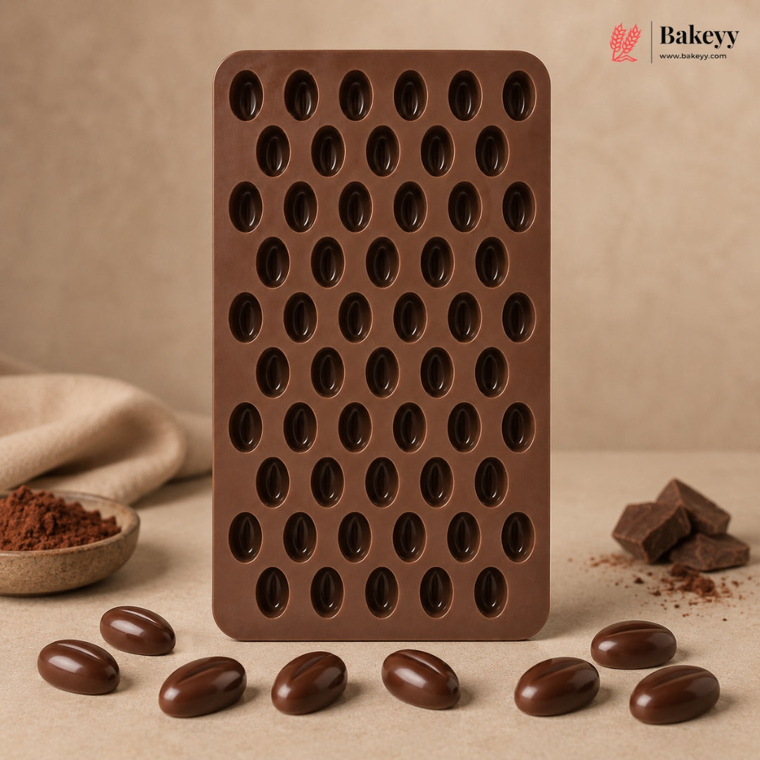 A brown silicone mold tray with multiple oval cavities, standing upright on a beige surface with chocolate-coated beans and chocolate pieces arranged around it. The Bakeyy logo appears in the top right corner.