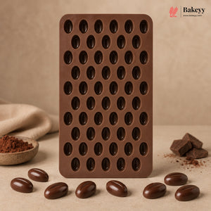 A brown silicone mold tray with multiple oval cavities, standing upright on a beige surface with chocolate-coated beans and chocolate pieces arranged around it. The Bakeyy logo appears in the top right corner.