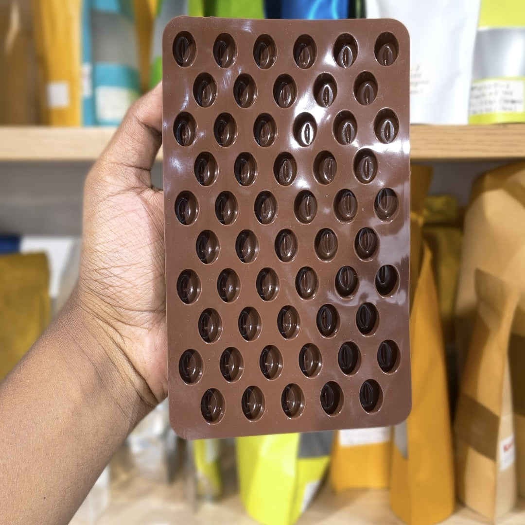 A hand holding a brown silicone mold tray with multiple oval cavities, set against a blurred background of shelves and packaged items.