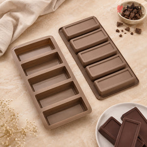 Two brown rectangular chocolate mold trays on a beige fabric surface, with a small bowl of chocolate chunks and chocolate bars used as styling props. No person or real environment is present, so this is Product/Packaging rather than Lifestyle.