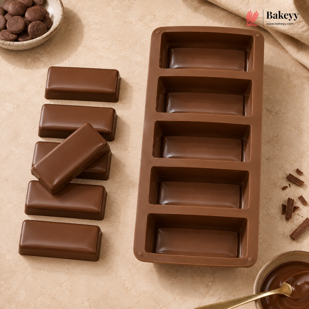 A brown silicone mold for making chocolate bars, shown next to five finished chocolate bars on a beige textured surface with baking props.