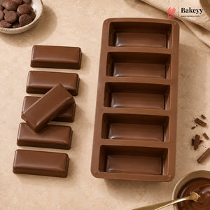 A brown silicone mold for making chocolate bars, shown next to five finished chocolate bars on a beige textured surface with baking props.