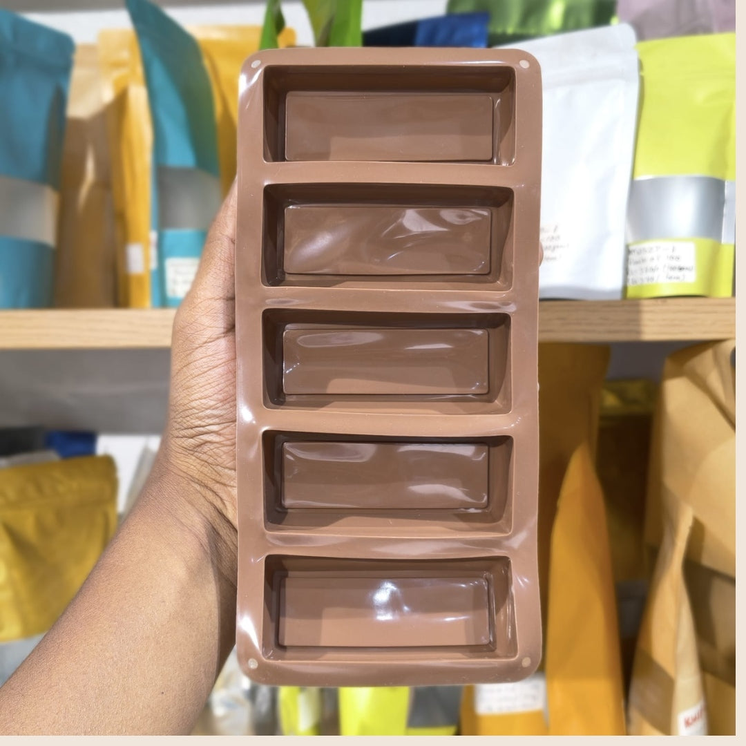 A hand holding a brown rectangular silicone mold with five elongated rectangular cavities, set against a blurred background of shelves with colorful pouches.