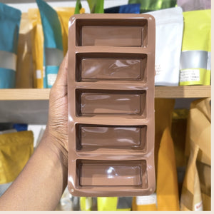 A hand holding a brown rectangular silicone mold with five elongated rectangular cavities, set against a blurred background of shelves with colorful pouches.
