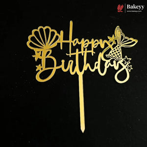 Gold metal 'Happy Birthday' cake topper with decorative seashell, butterfly, and star cutouts on a black background. A Bakeyy logo watermark is visible in the top right corner.