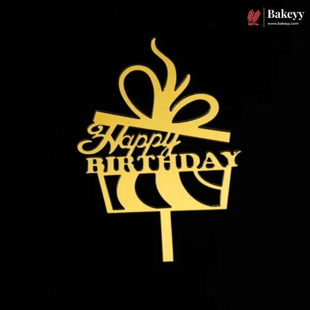 A gold metal cake topper shaped like a gift box with a bow and the words 'Happy Birthday' in script, shown on a black background. The Bakeey brand logo is visible in the top right corner.
