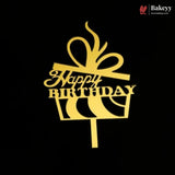A gold metal cake topper shaped like a gift box with a bow and the words 'Happy Birthday' in script, shown on a black background. The Bakeey brand logo is visible in the top right corner.