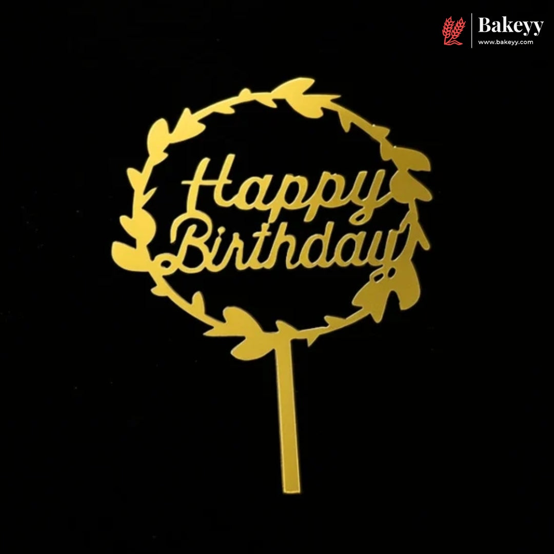 A gold metal cake topper with a circular leaf wreath and the words 'Happy Birthday' in script, shown on a black background. A Bakeey logo is visible in the top right corner.