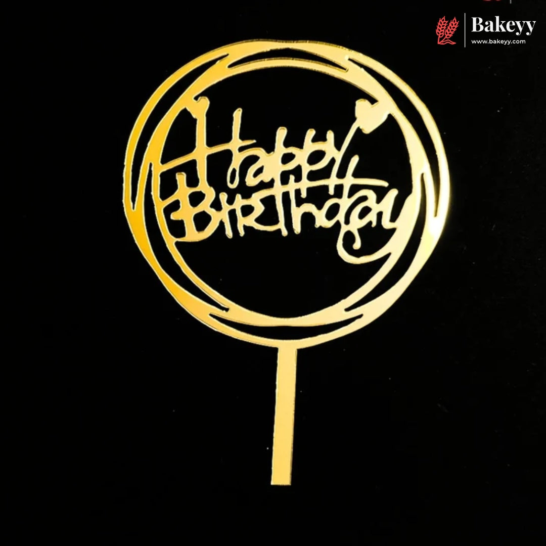 A gold-colored metal 'Happy Birthday' cake topper with a circular frame and stick, displayed on a solid black background. A Bakeey brand logo is visible in the top right corner.