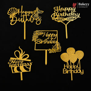 Six gold-colored plastic 'Happy Birthday' cake toppers in various designs, including a gift box, balloons, flowers, and a sign, arranged on a solid black background. A 'Bakeey' brand logo is visible in the top right corner.