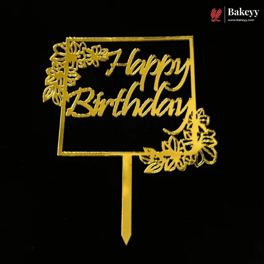 Gold-colored metal 'Happy Birthday' cake topper with floral accents and a stick, shown on a solid black background. A 'Bakeyy' brand logo is visible in the top right corner.