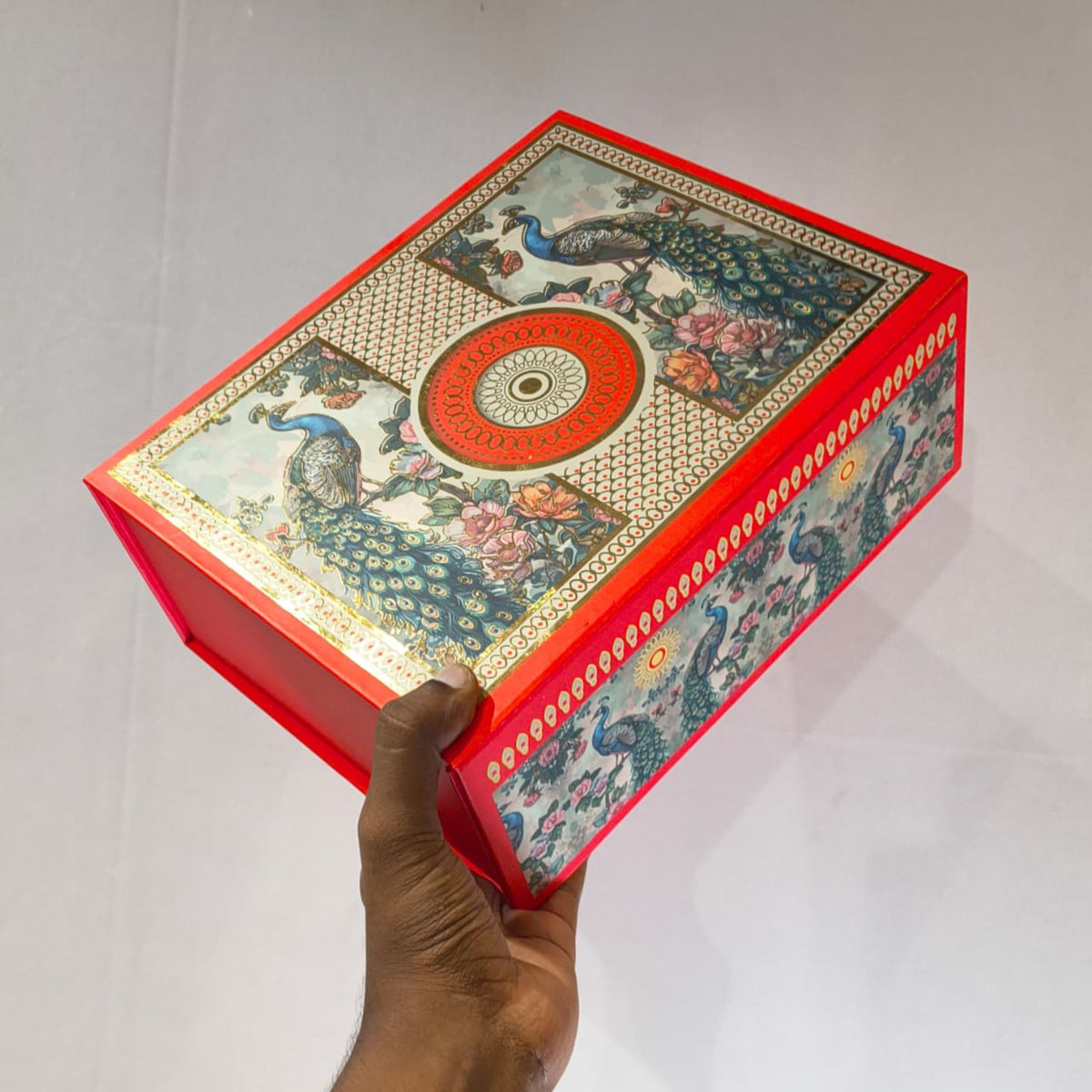 A hand holds a rectangular decorative box with a bright red border and an ornate peacock illustration on the lid and sides. The lid features a central circular motif and floral details, set against a plain light background.