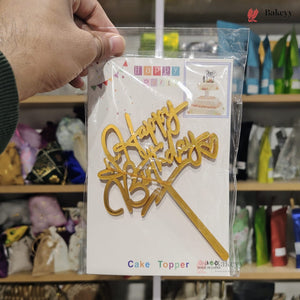 Gold 'Happy Birthday' cake topper in packaging held by a person with a blurred store background.